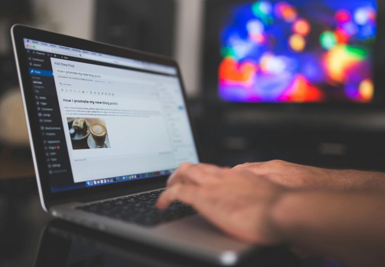 Hands typing on a laptop with a blog post visible, cozy indoor setting with colorful screen in background.